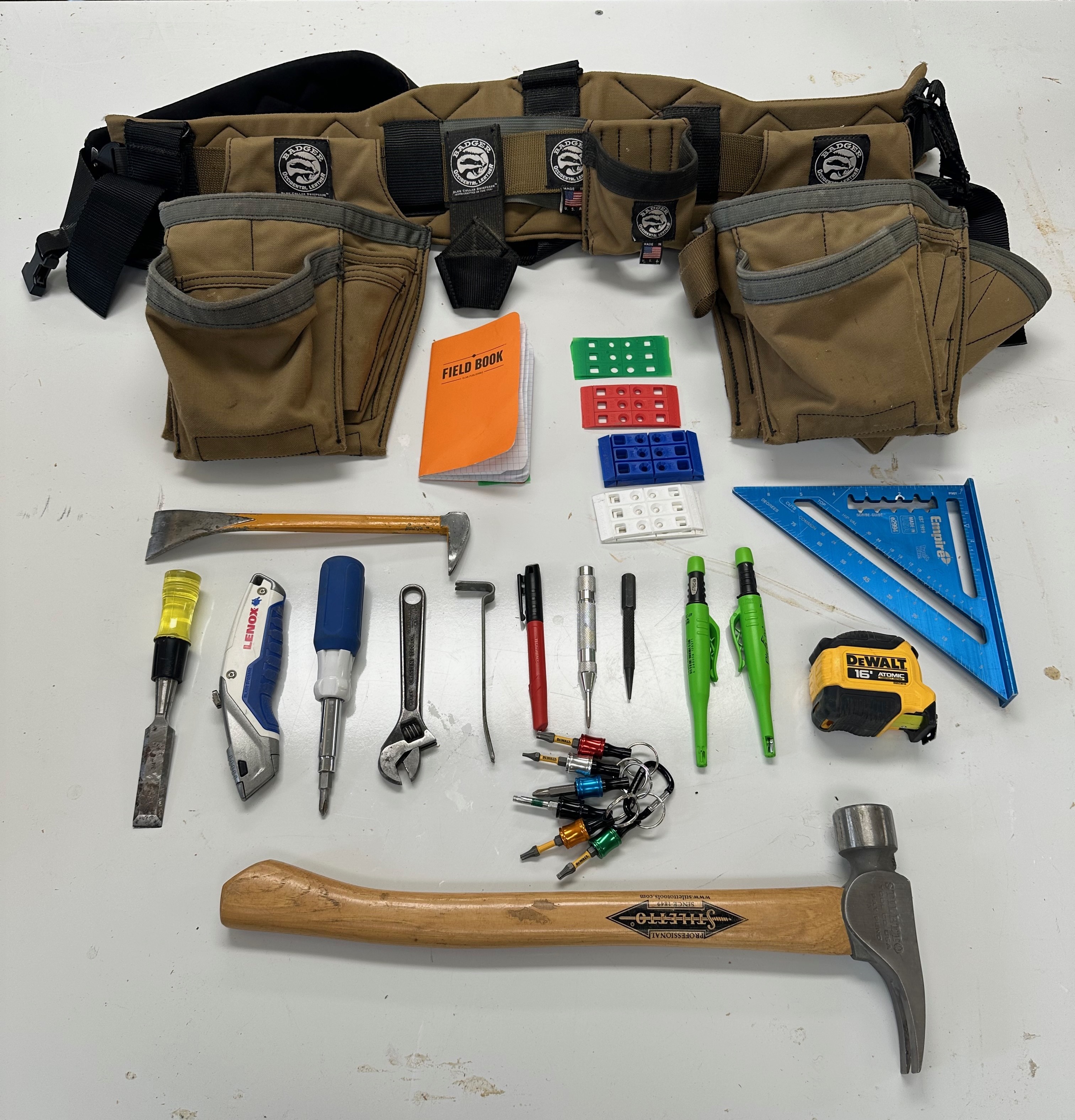 Badger tool belt above an organized layout of its notebooks, shims, square, marking tools, hand tools, driver bits, tape, and hammer.